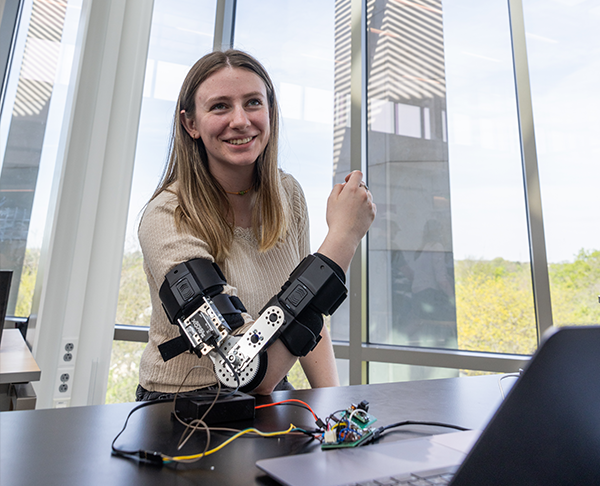 A female students smiles from the camera as she poses with a robotic arm attachment on her right arm.