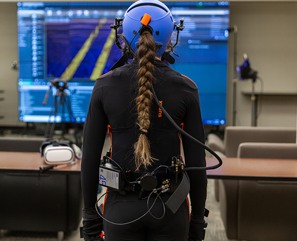 A student in a full, black body suit faces away from the camera. They are wearing a blue helmet that has wires coming from it and a large belt on their waist with battery packs and wires. 